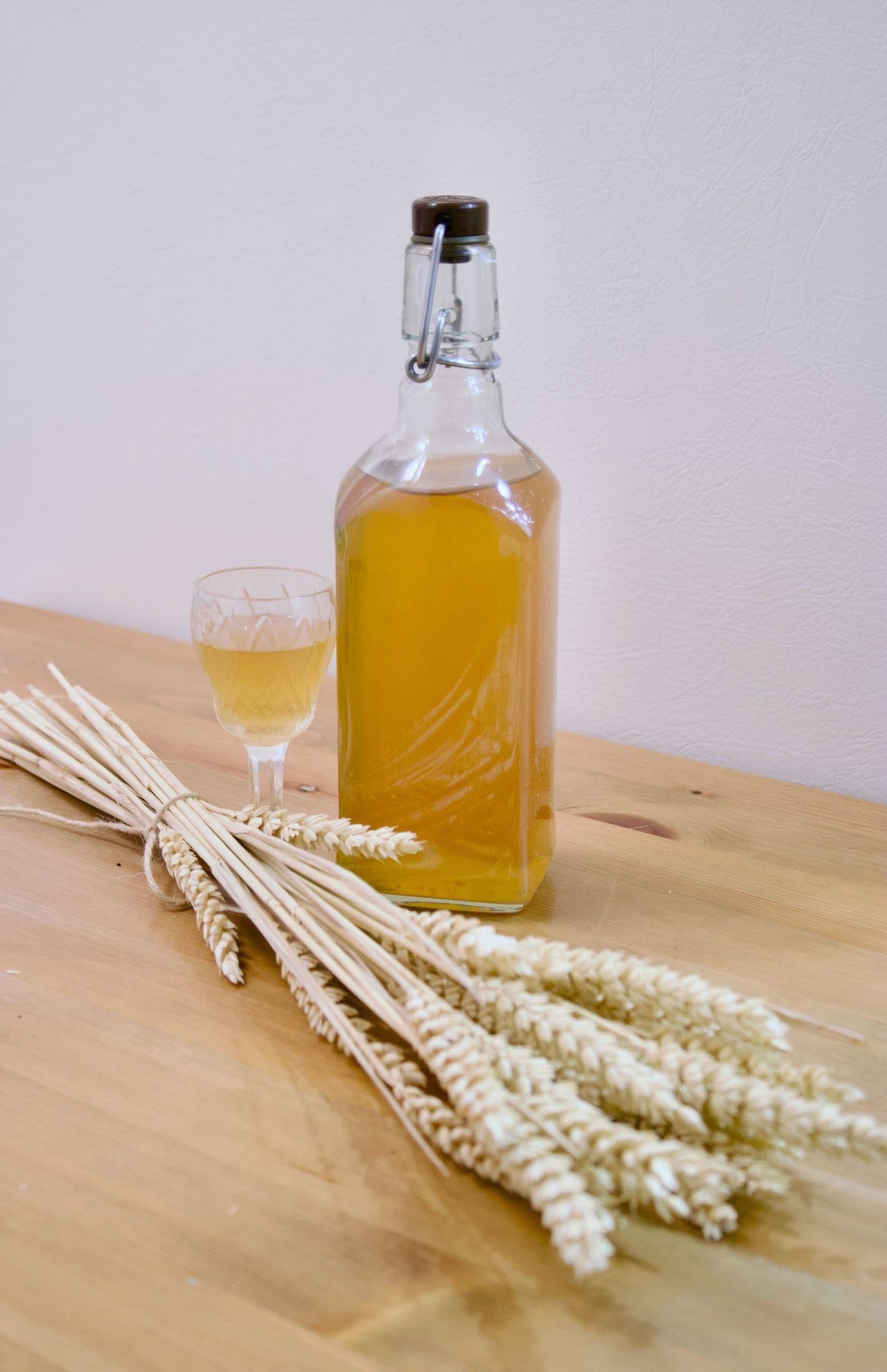 A swing-top bottle of amber botanical spirit beside a tasting glass and wheat stalks
